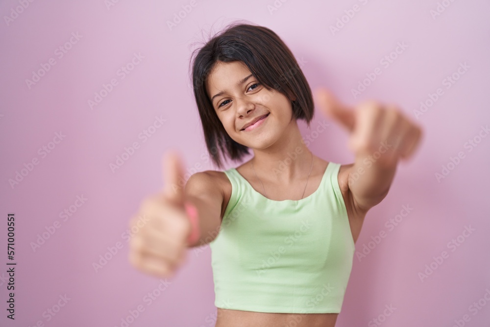Young girl standing over pink background approving doing positive gesture with hand, thumbs up smiling and happy for success. winner gesture. Young girl standing over pink background approving doing positive gesture with hand, thumbs up smiling and happy for success. winner gesture.