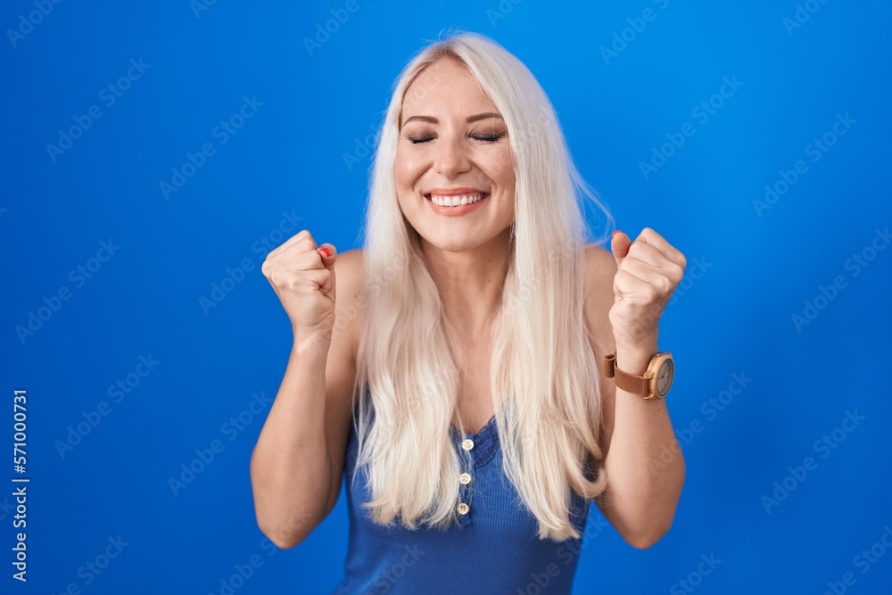 Fototapeta premium Caucasian woman standing over blue background excited for success with arms raised and eyes closed celebrating victory smiling. winner concept.