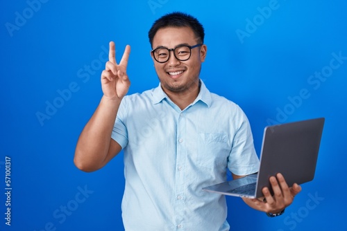 Chinese young man using computer laptop smiling with happy face winking at the camera doing victory sign with fingers. number two.