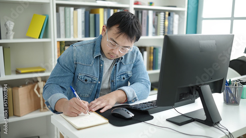 Young chinese man student using computer writing on notebook at library university