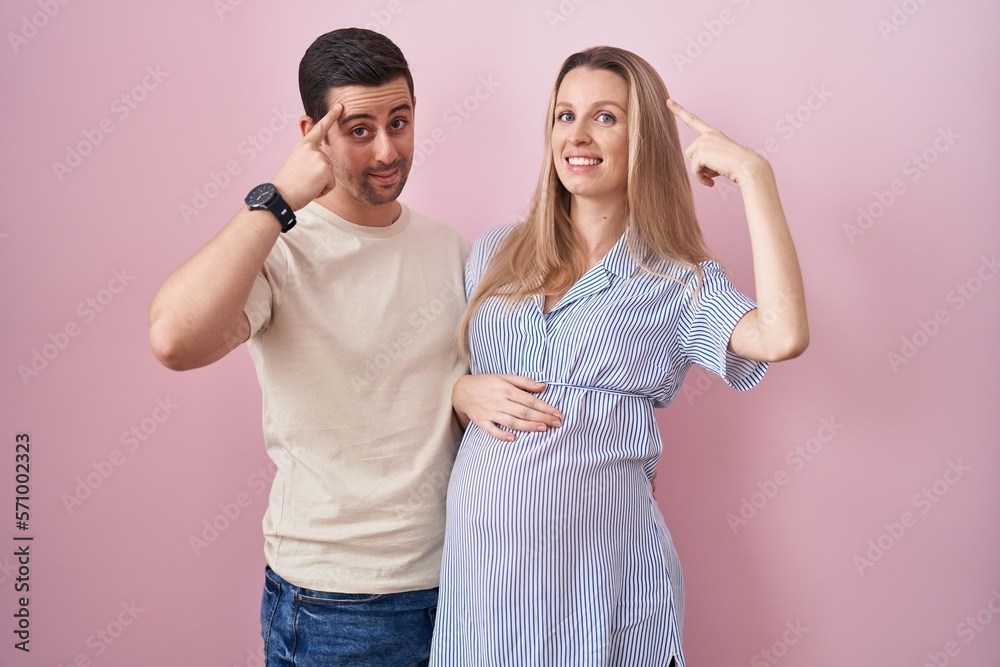 Young couple expecting a baby standing over pink background smiling pointing to head with one finger, great idea or thought, good memory Young couple expecting a baby standing over pink background smiling pointing to head with one finger, great idea or thought, good memory