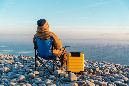 Bearded Man in brown jacket with yellow suitcase relaxing alone and sitting on beach chairs on the seaside  at sunrise. Travel Lifestyle concept © Iryna