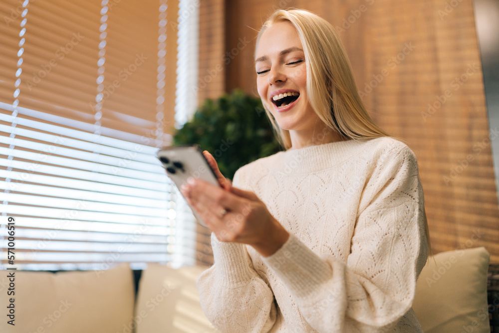 Low-angle view of cheerful young woman using mobile phone app enjoying online virtual chat, video call with friends in distance virtual meeting or recording stories for social media, sitting on sofa. Low-angle view of cheerful young woman using mobile phone app enjoying online virtual chat, video call with friends in distance virtual meeting or recording stories for social media, sitting on sofa.