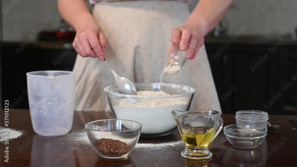 Woman prepares homemade bread with flax seeds. He gradually adds ingredients - salt, sugar, yeast, seeds. Mixes ingredients with flour in a transparent container. Home baking, food, traditions Woman prepares homemade bread with flax seeds. He gradually adds ingredients - salt, sugar, yeast, seeds. Mixes ingredients with flour in a transparent container. Home baking, food, traditions