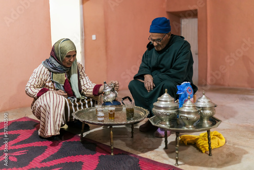 Berber man and woman with traditional clothes in the living room of their house preparing tea for their guests with silver utensils