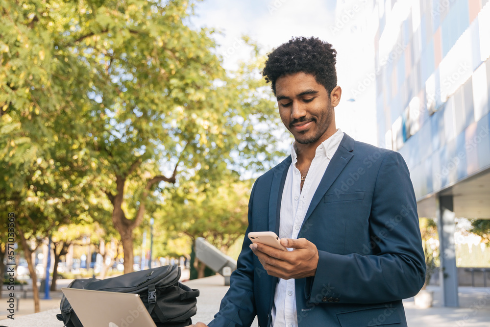 Black businessman in suit working outdoors with laptop during a break looking at his mobile phone and smiling Black businessman in suit working outdoors with laptop during a break looking at his mobile phone and smiling
