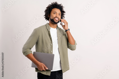 Busy handsome confident bearded african man in casual wear talking on smartphone carrying laptop isolated on white, male entrepreneur with curly afro airstyle taking a call on the mobile phone photo