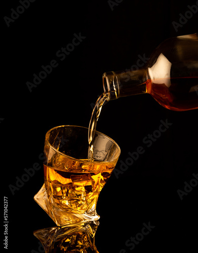 Pouring alcohol and throwing an ice cube into a glass with splashes on a black background