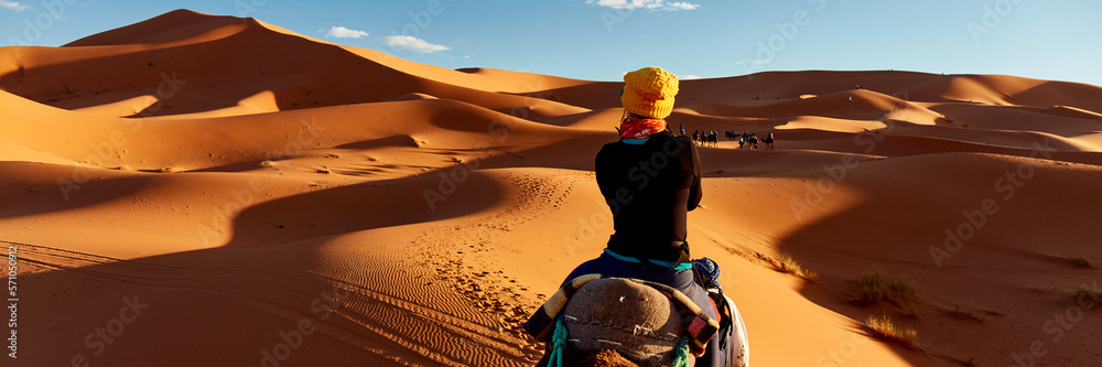 A young women in a yellow cap rides a camel  through the dunes in the Sahara Desert. View of the woman from behind, in the background, small silhouettes of other tourists. Merzouga, Morocco - obrazy, fototapety, plakaty 