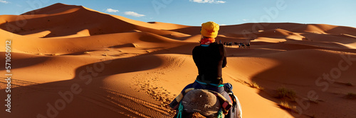 A young women in a yellow cap rides a camel through the dunes in the Sahara Desert. View of the woman from behind, in the background, small silhouettes of other tourists. Merzouga, Morocco