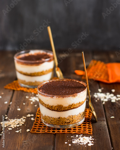 Postre en vaso a base de capas de copos de avena, crema de queso y cacao en polvo, posavasos naranja y mesa de madera photo