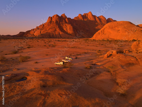 Aerial view of campsite at the foot of Spitzkoppe. Three offroad camper cars on campsite against red, bald granite peaks illuminated by setting sun. Travelling Namib desert, Namibia. 