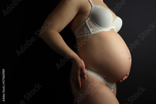 Pregnant woman in underwear. She is 30 weeks pregnant and has a big belly. Expecting a child. Third pregnancy. Pregnant girl. Black background and white underwear