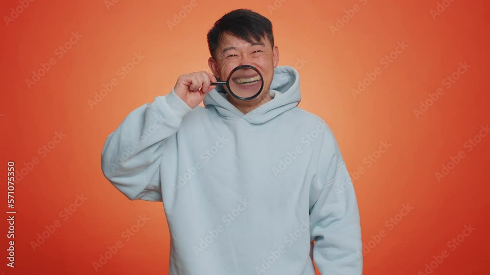 Asian man holding magnifier glass on healthy white teeth, looking at camera with happy expression, showing funny silly face smiling mouth. Handsome chinese guy isolated on orange studio background Asian man holding magnifier glass on healthy white teeth, looking at camera with happy expression, showing funny silly face smiling mouth. Handsome chinese guy isolated on orange studio background
