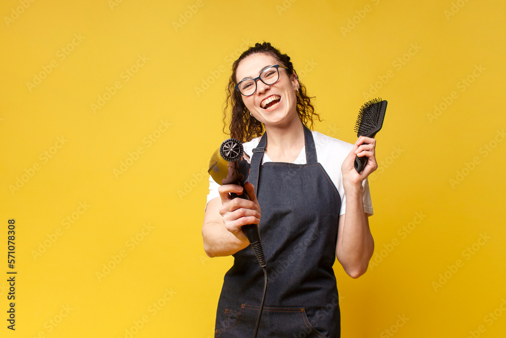 young girl hairdresser in uniform holds hair dryer and comb and smiles ...