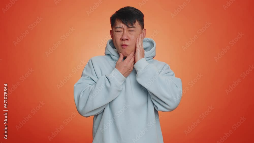 Asian man touching sore cheek suffering from toothache cavities or gingivitis waiting for dentist appointment gums disease. Handsome chinese guy indoors studio shot isolated on orange background