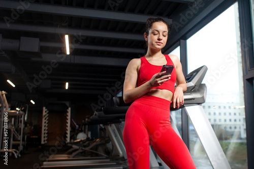 young sporty girl in red sportswear uses smartphone in the gym, attractive slim woman holds the phone in training