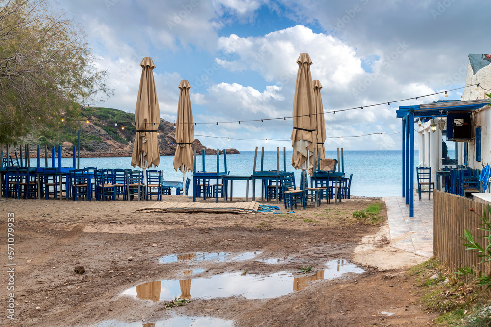 A seaside, beachfront outdoor cafe with blue tables and chairs stacked up, on an overcast Autumn day along the coast of Cape Sounion, Greece.