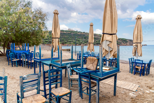 Fototapeta Naklejka Na Ścianę i Meble -  A seaside outdoor cafe with blue tables and chairs stacked up, on an overcast Autumn day along the coast of Cape Sounion, Greece, with the ancient Temple of Poseidon