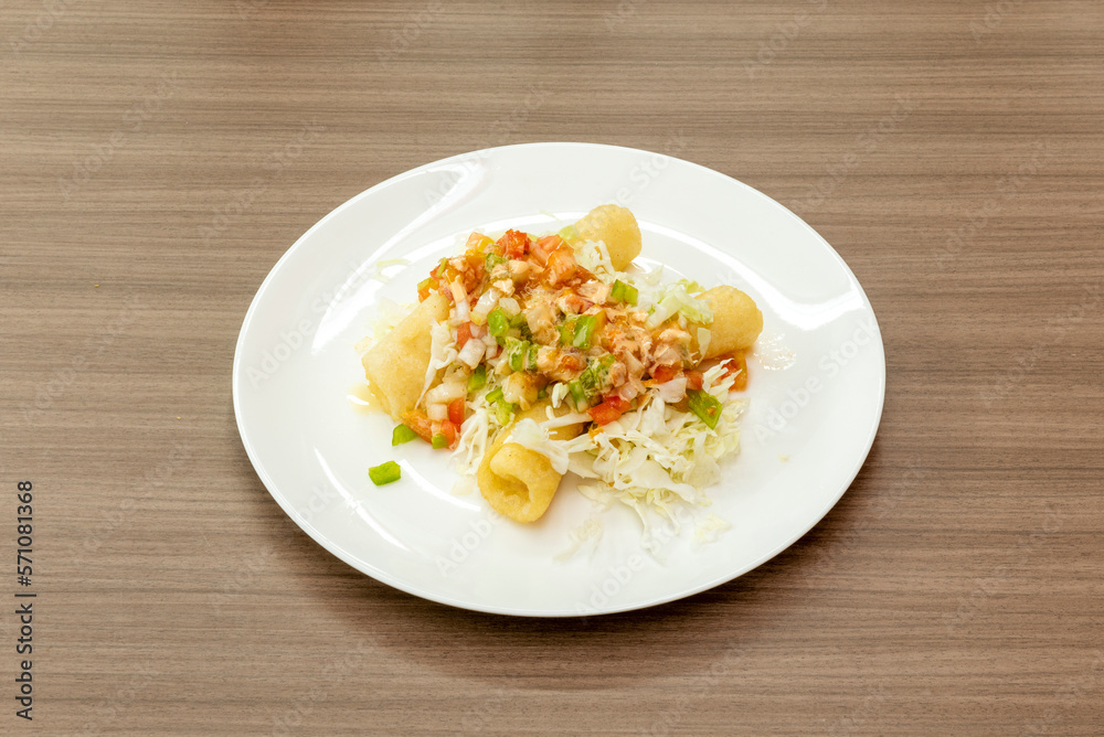 A bright white plate of Mexican flautas with minced meat stew and salad on a light brown table A bright white plate of Mexican flautas with minced meat stew and salad on a light brown table