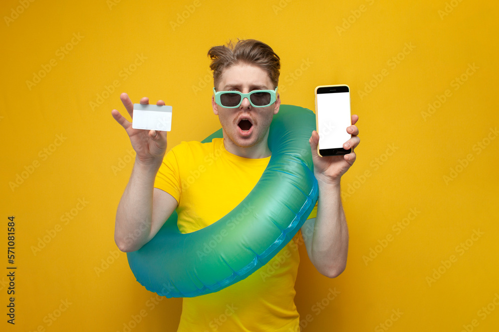 young guy tourist in the summer on vacation holds a phone and a bank card and shows surprise