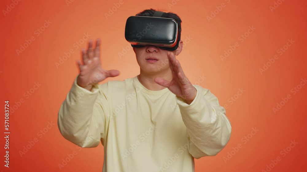Asian man using headset helmet to play simulation game app online. Watching virtual reality 3D 360 video content. One guy in VR goggles isolated on studio orange background. Futuristic technology Asian man using headset helmet to play simulation game app online. Watching virtual reality 3D 360 video content. One guy in VR goggles isolated on studio orange background. Futuristic technology