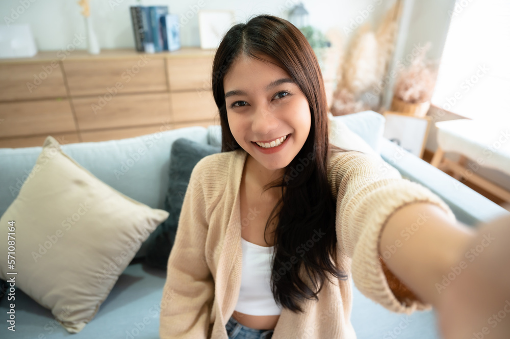 Pretty young asian female with big smile sitting at living room. She having fun taking light cheerful selfie on blurred background