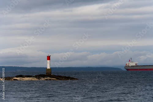 An image of a small light house in the pacific ocean with a cargo ship in the distance. 