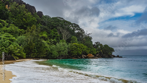 The waves of the turquoise ocean roll onto the shore of a tropical island. Foam spreads over the sand. Boulders in the water. A hill overgrown with green vegetation, against a blue sky and clouds