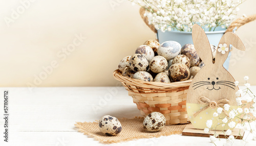 Woven basket of quail eggs, wooden bunny, white flowers on white wooden table on beige Easter card photo