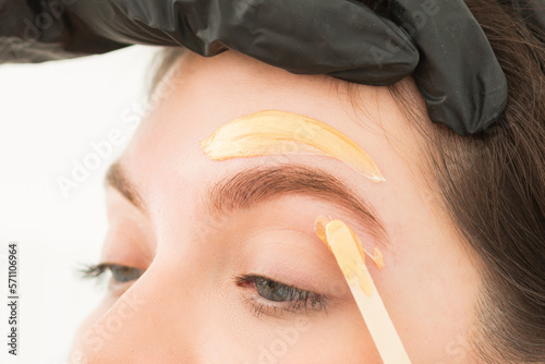 Young woman undergoing eyebrow correction procedure, waxing, wax depilation of eyebrow hair, brow correction in beauty salon, closeup. Golden wax and wooden sticks, tools. Eyebrow master