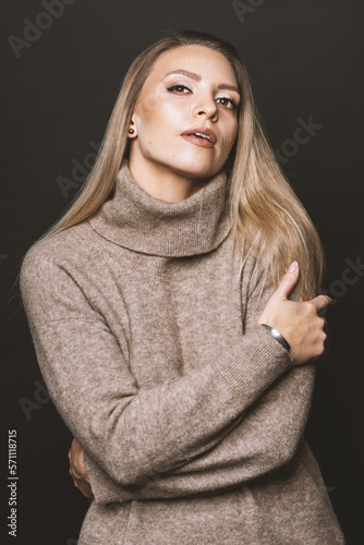 Studio portrait of beautiful brunette woman with day make-up and cozy beige sweater. Model hugging herself with hands and looking at the camera. Toned image with beige color. Soft filter applied