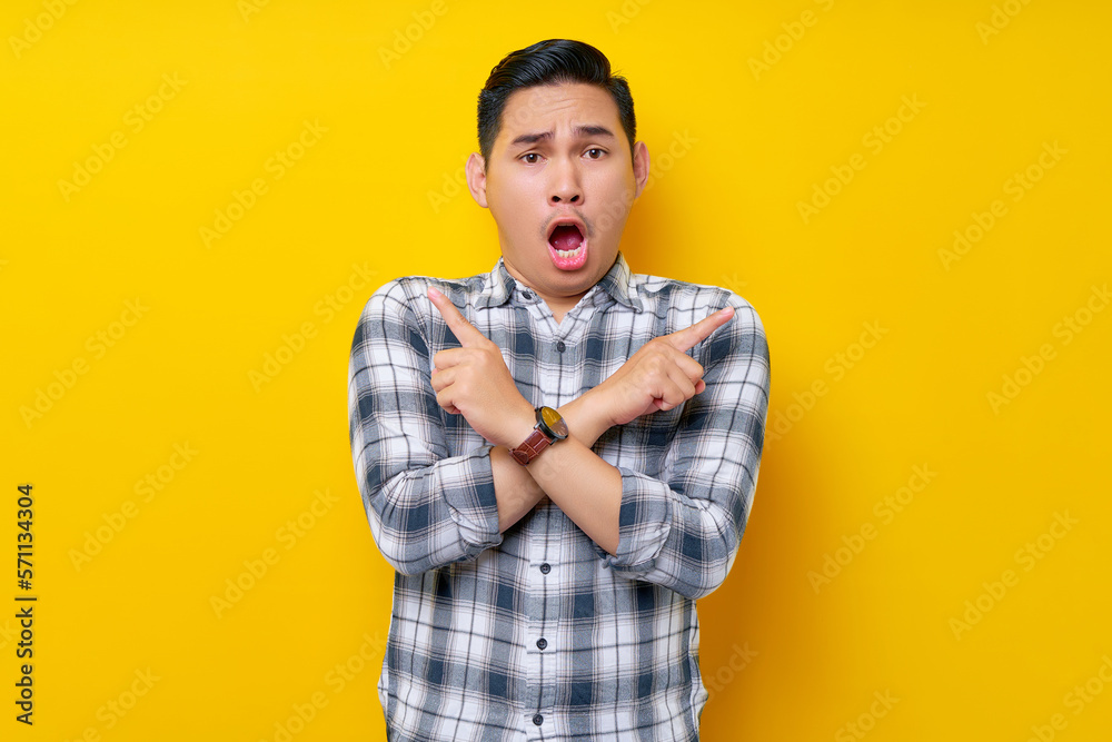 Shocked Young Asian man in casual clothes crossed arms and points sideways, chooses between two options, indicates left and right isolated on yellow background. People lifestyle concept Shocked Young Asian man in casual clothes crossed arms and points sideways, chooses between two options, indicates left and right isolated on yellow background. People lifestyle concept