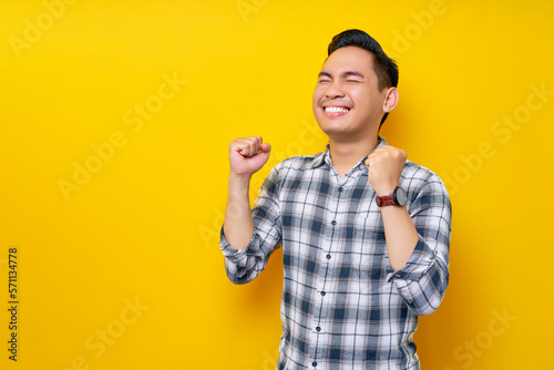 Excited young handsome Asian man celebrating victory and raised fists isolated on yellow background. People lifestyle concept