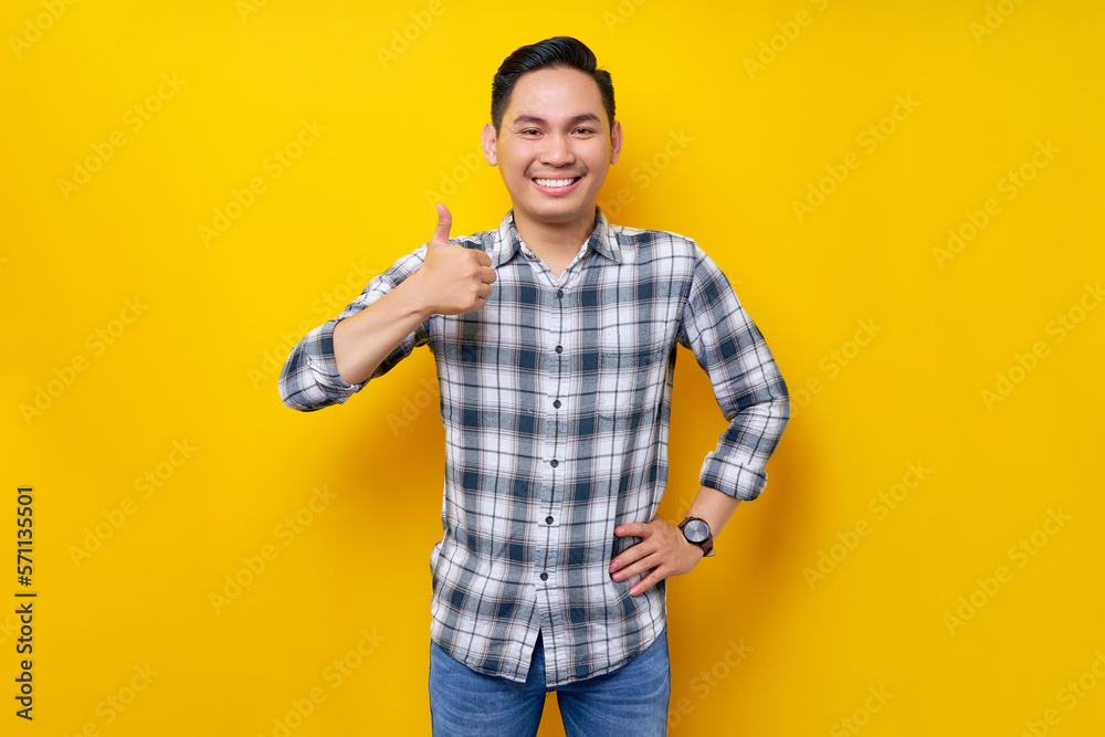 Smiling young handsome Asian man wearing casual clothes showing thumb up gesture, looking at camera isolated on yellow background. People lifestyle concept
