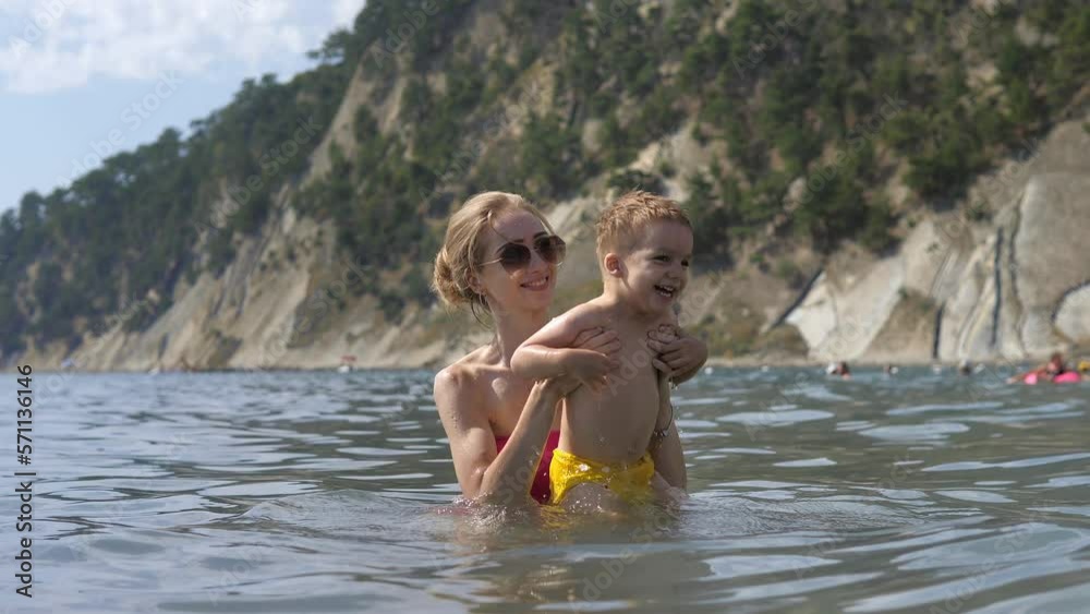 Young mother in sunglasses is playing with 2 years old boy in seawater sunny day outdoor on natural rock background. Happy people have fun on summer beach holiday. Young mother with son relax at sea. Young mother in sunglasses is playing with 2 years old boy in seawater sunny day outdoor on natural rock background. Happy people have fun on summer beach holiday. Young mother with son relax at sea.