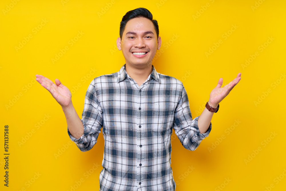Smiling young handsome Asian man wearing a plaid shirt Standing spreading hands, having fun looking at the camera isolated on yellow background. People lifestyle concept Smiling young handsome Asian man wearing a plaid shirt Standing spreading hands, having fun looking at the camera isolated on yellow background. People lifestyle concept