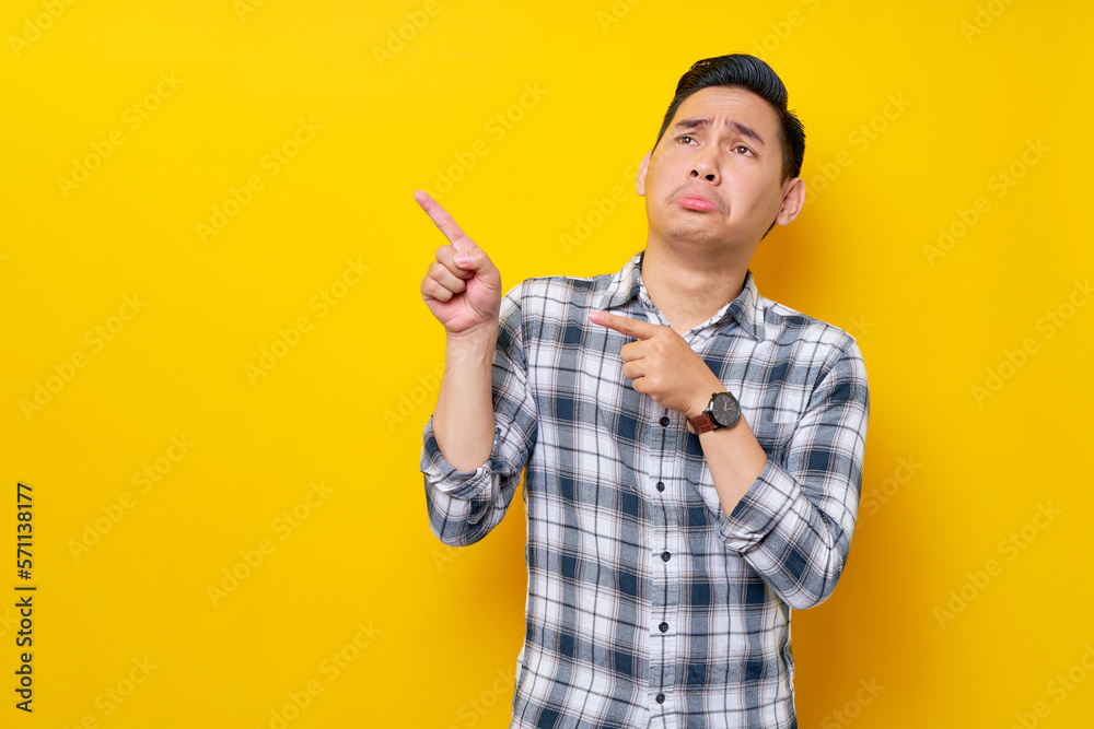 Confused young handsome Asian man 20s wearing a plaid shirt pointing a finger at the advertising area isolated on yellow background. People lifestyle concept Confused young handsome Asian man 20s wearing a plaid shirt pointing a finger at the advertising area isolated on yellow background. People lifestyle concept