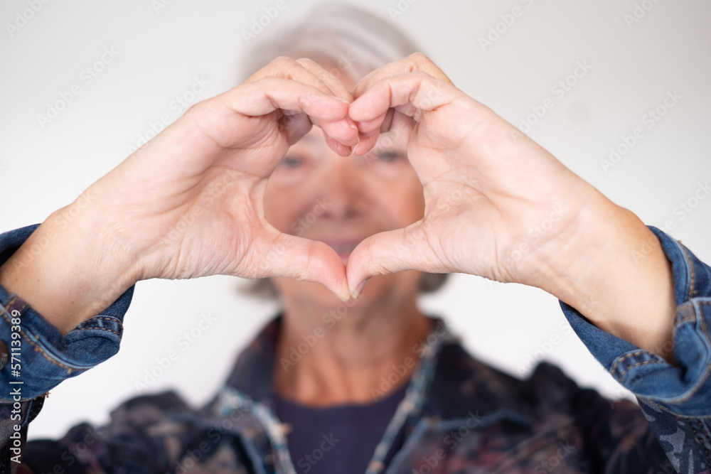 Blurred senior woman in denim jacket looking at camera making heart shape with hands, isolated on white background. Care, love, emotion, feelings, body language Blurred senior woman in denim jacket looking at camera making heart shape with hands, isolated on white background. Care, love, emotion, feelings, body language