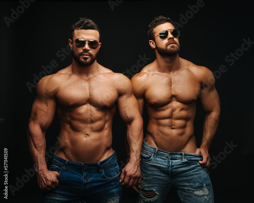 Two shirtless hunks at black background. Fitness models couple in jeans and sunglasses. Two muscular handsome men with six pack abs in studio.