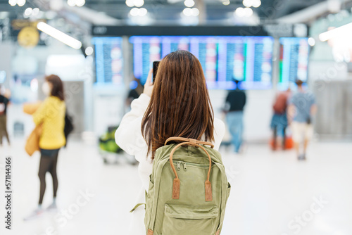 Young woman with backpack looking at the flight time information board  and using smartphone in international airport. Travel, Vacation, trip and Transport concept © Jo Panuwat D
