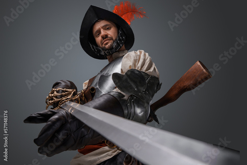 фотографія Isolated on grey medieval musketeer staring at camera and pointing sword to camera