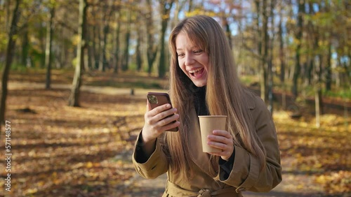 Front view of positive, happy lady standing in park, using smartphone, scrolling. Attractive woman wearing stylish trech, holding paper cup, drinking coffee. Concept of modern lifestyle. photo