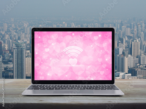 Heart love wifi flat icon modern laptop computer with pink screen on wooden table over city tower and skyscraper, Internet online love connection, Valentines day concept