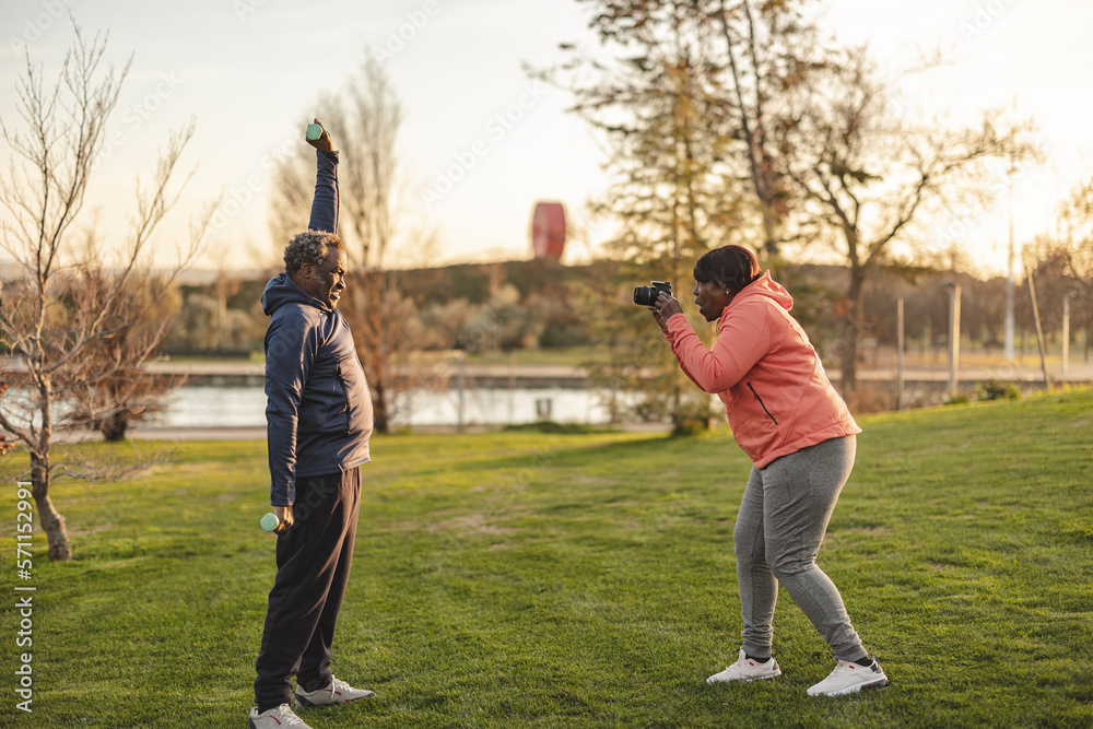 Senior woman clicking photos of man lifting dumbbells at park Senior woman clicking photos of man lifting dumbbells at park