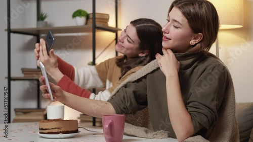 Network addiction. Selfie women. Home leisure. Happy coquettish female friends sitting desk with cake making selfie on personal smartphone light room interior.