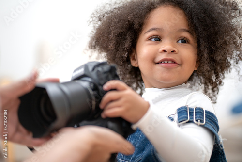 Cute little girl holding camera with smile. Portrait of playful multiracial kid interested in photography. Beautiful curly hair baby child playing with camera and lens in father hand. Family enjoyment © Nassorn