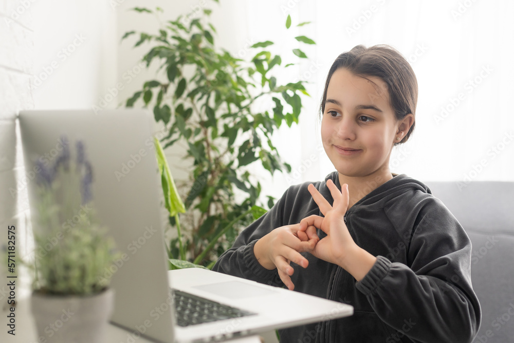Distant education. Friendly teen girl having online lesson via laptop and showing gesturing in sign language, studying online from home Distant education. Friendly teen girl having online lesson via laptop and showing gesturing in sign language, studying online from home