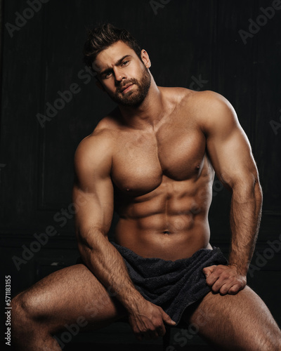Brutal sexy man with six pack abs in towel at black background. Handsome fitness male model sitting in studio. Bearded muscled guy covering with towel in studio.