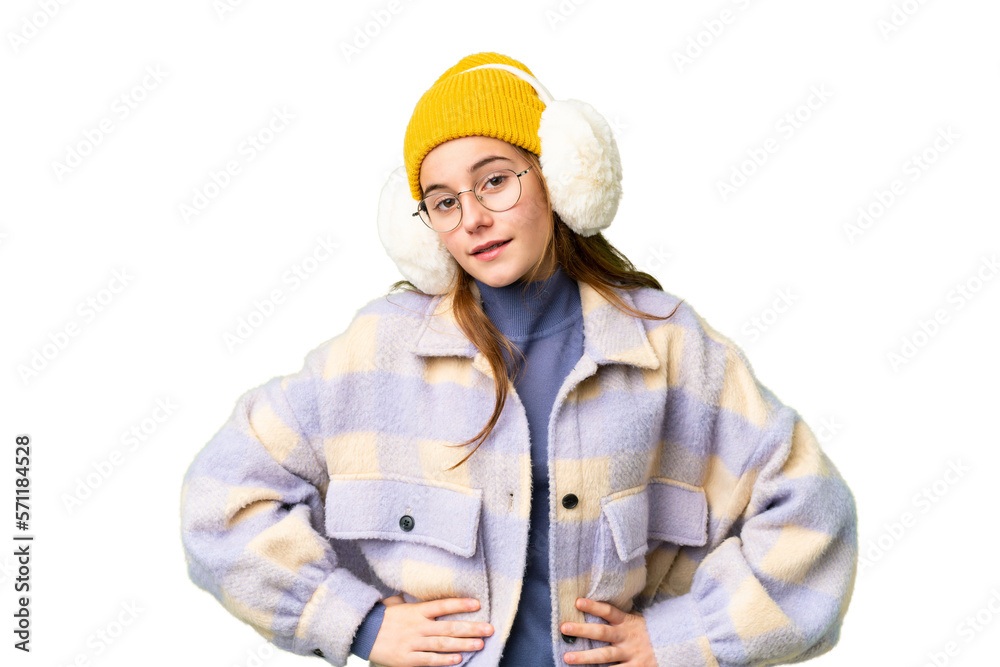 Teenager girl wearing winter muffs over isolated chroma key background posing with arms at hip and smiling Teenager girl wearing winter muffs over isolated chroma key background posing with arms at hip and smiling